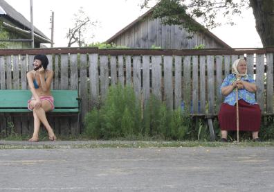 Deux femmes sont assises sur deux bancs a l'extrémité du cadre de la photo, devant une clôture. A gauche une jeune femme en maillot de bain et à droite une vieille dame avec une canne