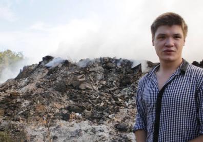 Un jeune homme en chemise se tient en premier plan devant une foret et un tas de gravas entrain de bruler