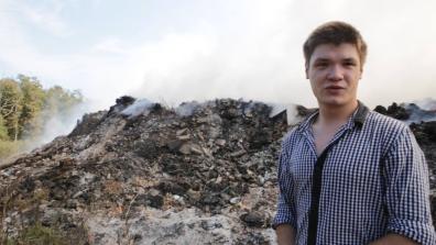 Un jeune homme en chemise se tient en premier plan devant une foret et un tas de gravas entrain de bruler