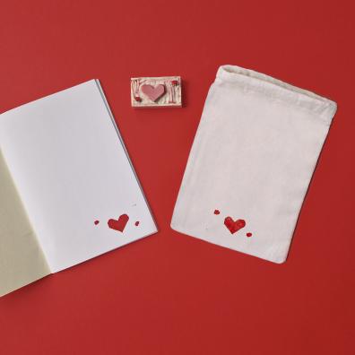 un carnet blanc ouvert et une pochette blanche à côté avec un petit coeur rouge dessiné. Au milieu des deux objets il y a une tampon de la forme du coeur