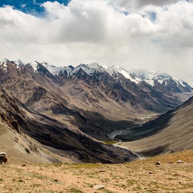 Vue entre deux chaînes de montagnes, un ruisseau au milieu, les pics de montagnes enneigés et un ciel blanc de nuages 