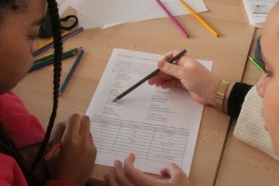 Two students work at a desk and write on a sheet of paper.