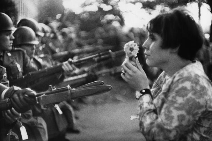 Photo en noir et blanc : des soldats pointent leurs armes sur une jeune femme qui se tient droite avec une fleur devant le visage pour se protéger 