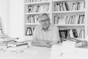 Portrait d'un homme à son bureau, il porte des lunettes et une chemise. Il est assis devant une bibliothèque