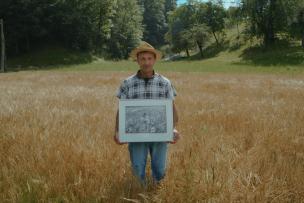 Un homme dans un champ de blé portant un chapeau et une chemise à carreaux. Il tient dans ses mains un cadre avec une photo