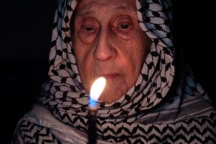 Portrait d'une vieille dame portant un keffieh palestinien blanc dans l'obscurité, tenant une lampe.